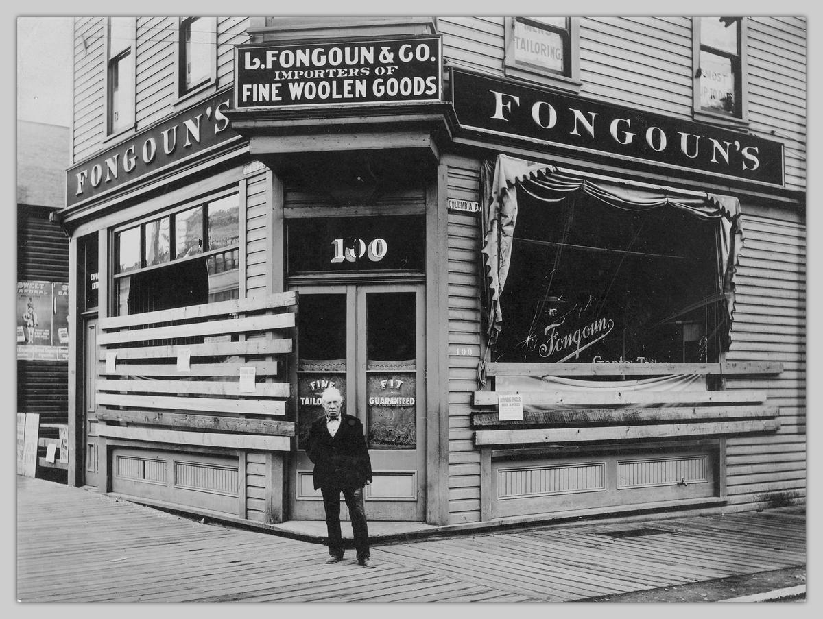 Black and white photograph of a damaged business with boarded up windows and a man standing at the front.