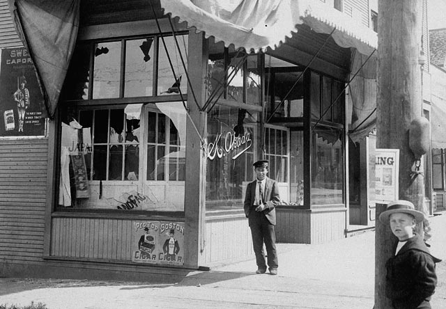 A store with broken windows, with a man standing in front of it.