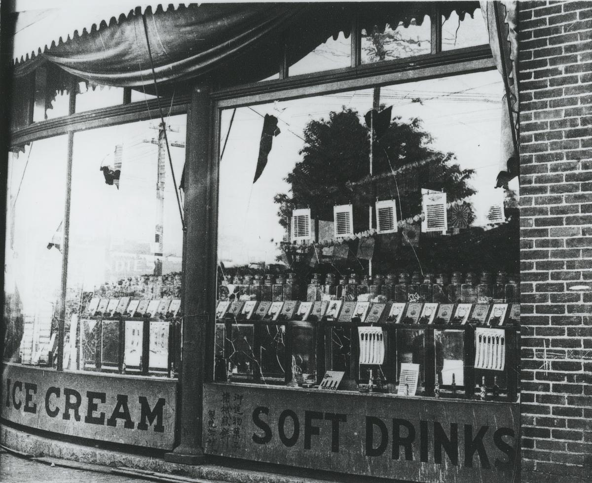 Black and white photograph of an ice cream and soft drinks store with broken windows.