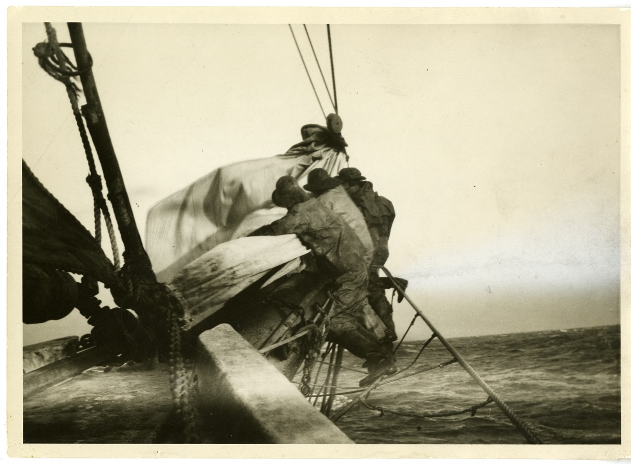 Stowing a Jib Black-and-white photo of three fishermen in oilskins and hats leaning out over the bow, hauling in a large, flapping sail. Rough sea stretches to the horizon behind them.