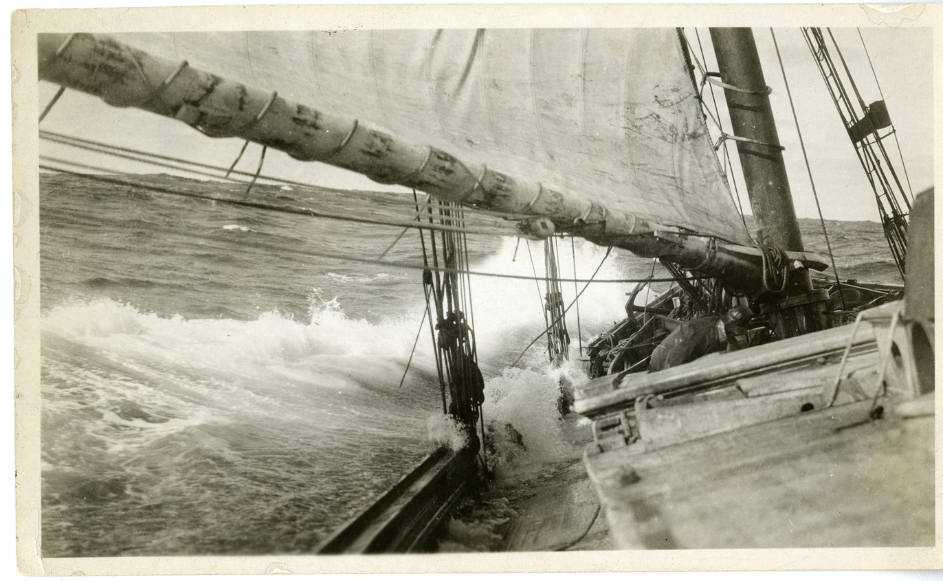 Albert J. Lutz Black-and-white photo looking along the deck of a schooner as a wave breaks over its side. A large sail and boom stretch overhead, and a fisherman in oilskins crouches near the mast.