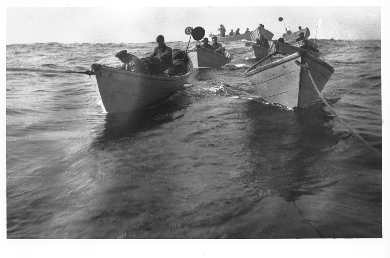 Flying Set Black-and-white photo of several small wooden boats being towed in a line across open water. Fishermen sit in each boat, and some boats carry tall marker poles with flags.