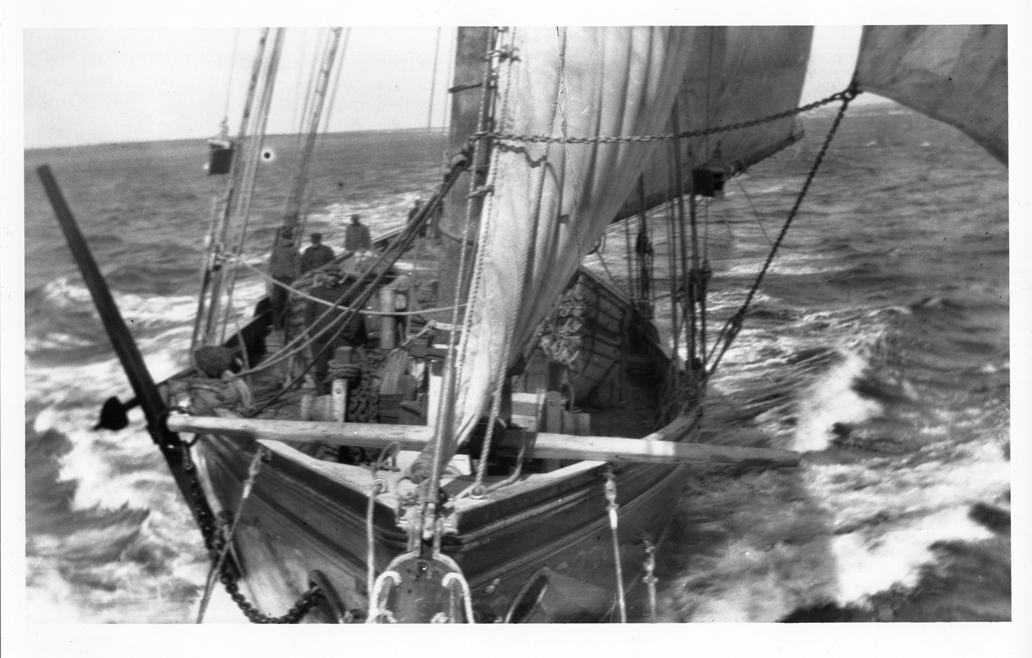 Schooner D.M. Smart Black-and-white photo looking down from the bowsprit onto the bow and deck of a schooner. Its sails are full of wind, and a few crew members stand on deck as the ship cuts through choppy water.