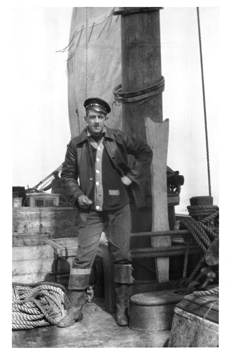 Frederick William Wallace Black-and-white photo of a young man in a peaked cap, jacket and tall rubber boots. He stands on deck beside the mast with one hand on his hip, with coils of rope at his feet.