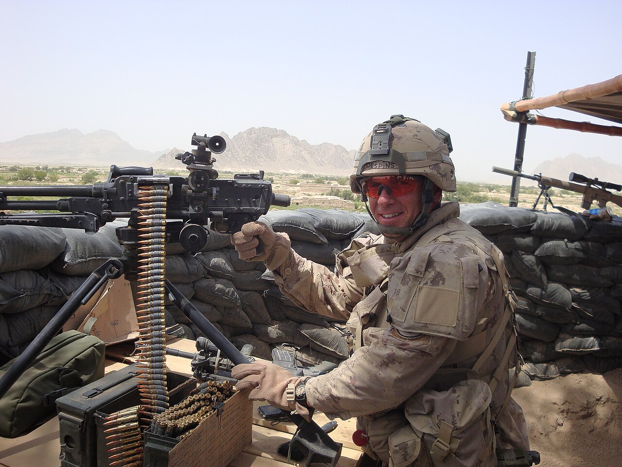 Un soldat canadien à côté d'une mitrailleuse C6 montée sur trépied à Kandahar, en Afghanistan (2009) Un soldat souriant tient une C6 par la poignée pistolet et se tient près de sacs de sable.