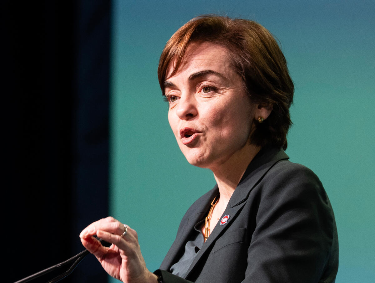 A woman with short hair wearing a dark blazer speaking at a microphone.