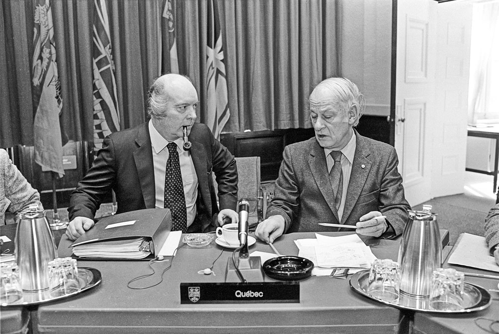 Claude Morin et René Lévesque Des hommes assis à une table, avec des drapeaux en arrière-plan.