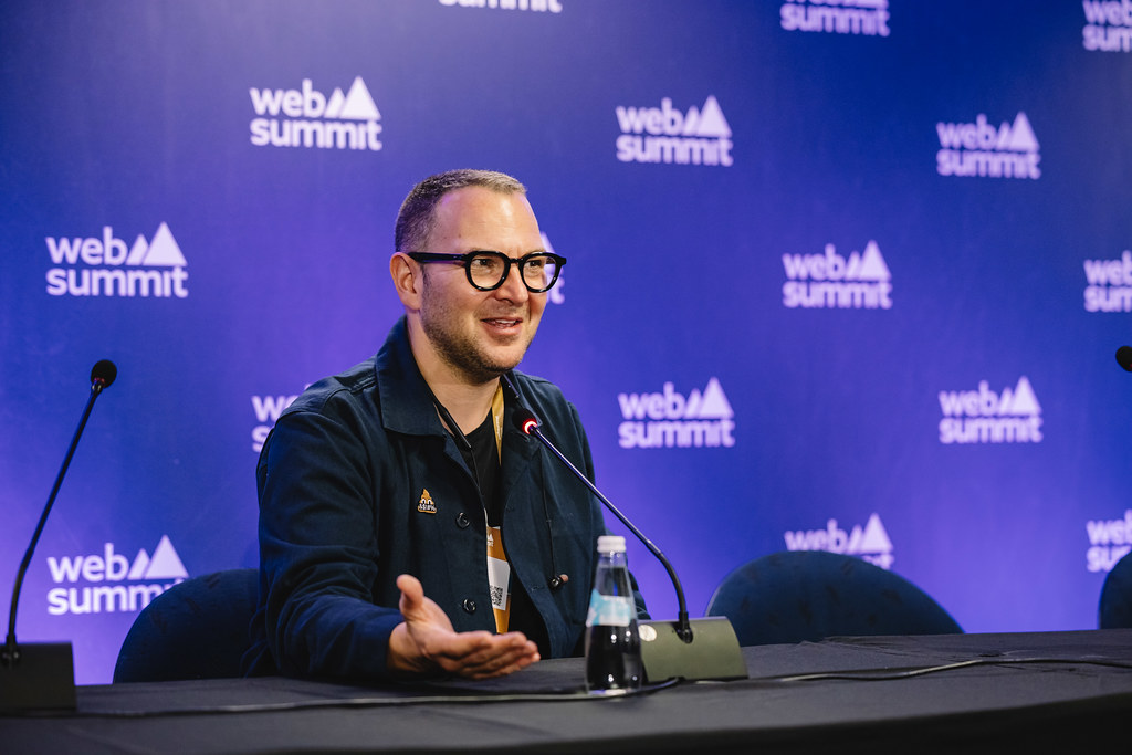 Un homme assis à une table avec un micro. Il y a un mur violet avec des inscriptions de Web Summit.
