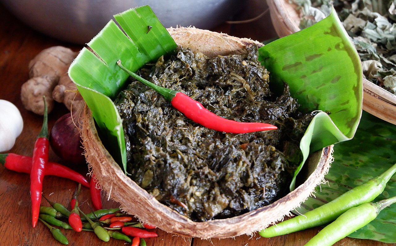 A coconut with a green leaf holding a black-coloured food with a chilli on top.