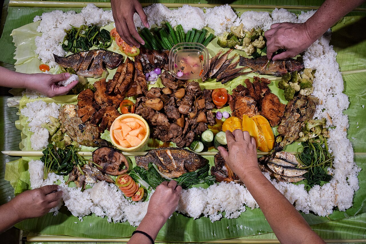 Several dishes on top of rice and leaves underneath. The hands of several people serving themselves.