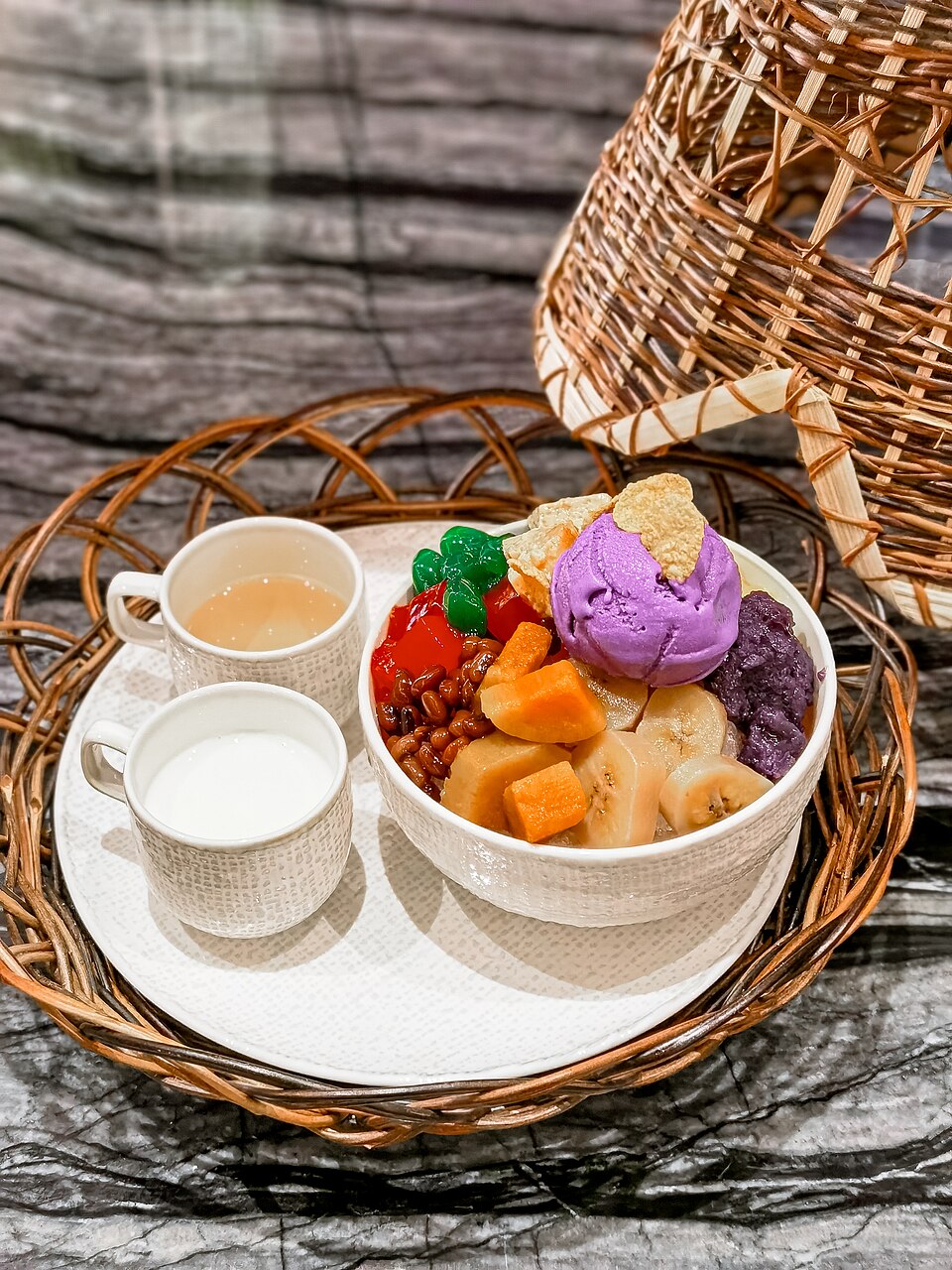 A small bowl full of fruits, beans and various other deserts, topped with purple yam ice cream. Two cups with liquids are seen to the left.