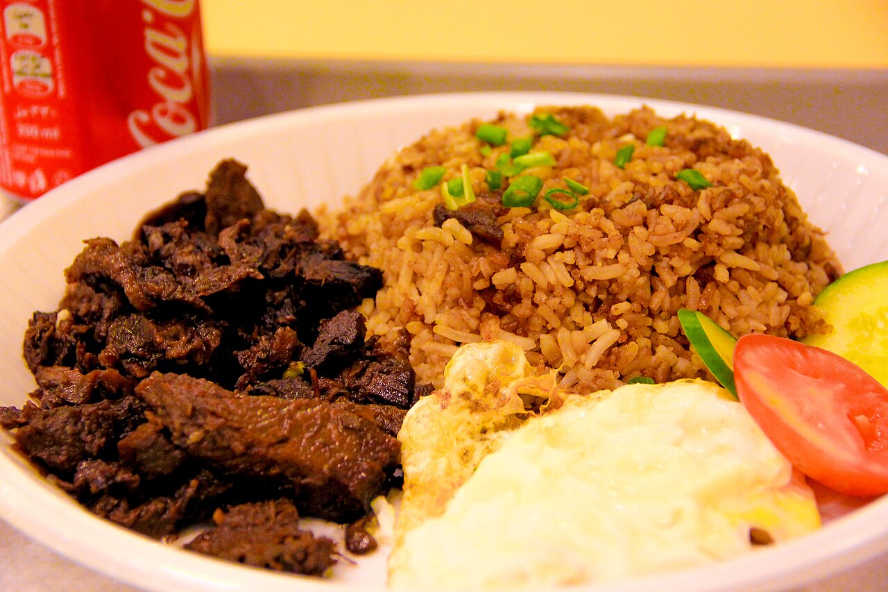 A bowl with rice, meat, an egg, and vegetables. A red can is seen behind.