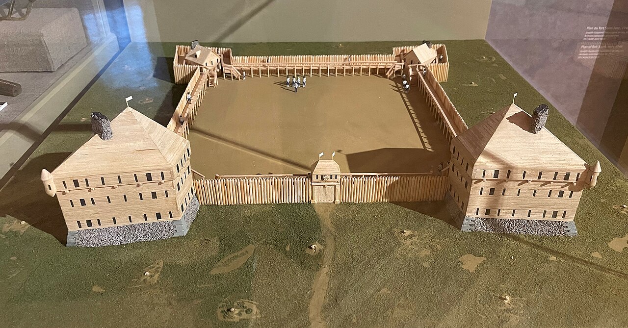 Wooden scale model on fake grass behind a glass pane.