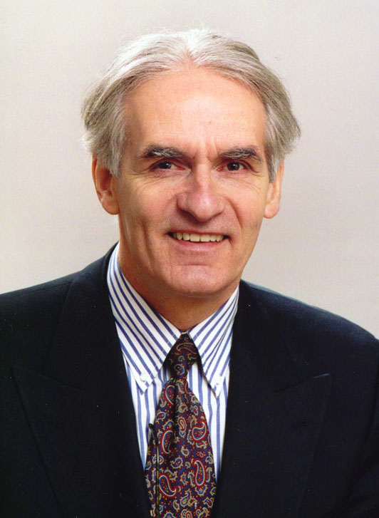 A smiling white man. He is wearing a dark suit, a tie, and a white shirt with blue stripes.