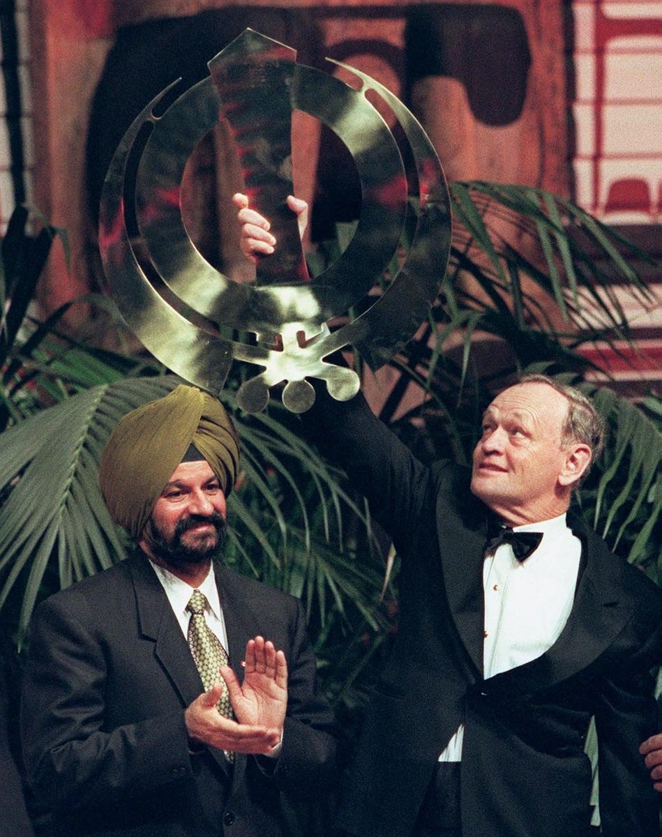 A man wearing a dark yellow turban clapping while a white man on the right holds up a Sikh symbol