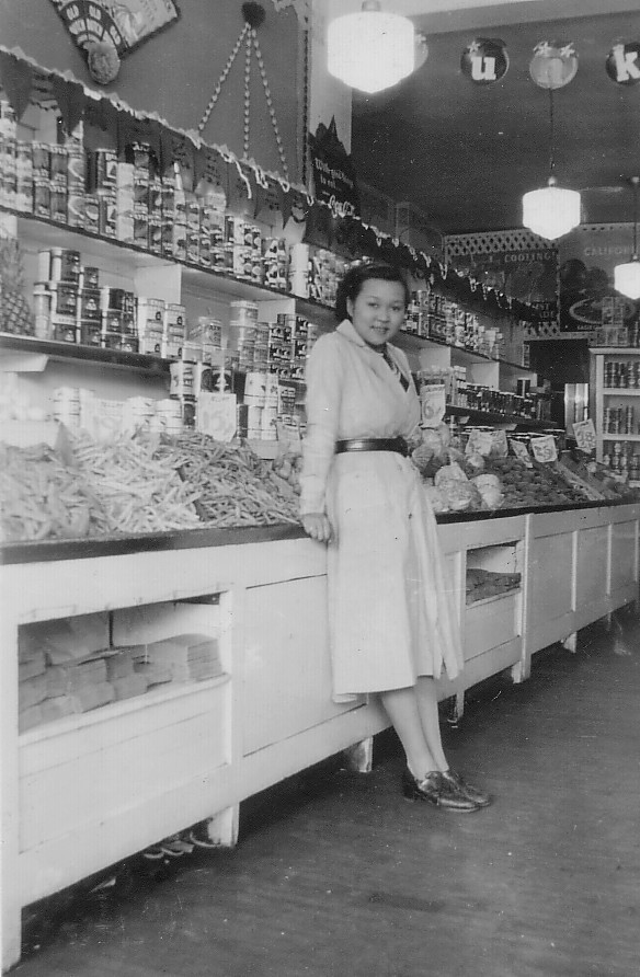 Jean Lumb at Her Fruit Store Woman leaning against a row of produce in a store.