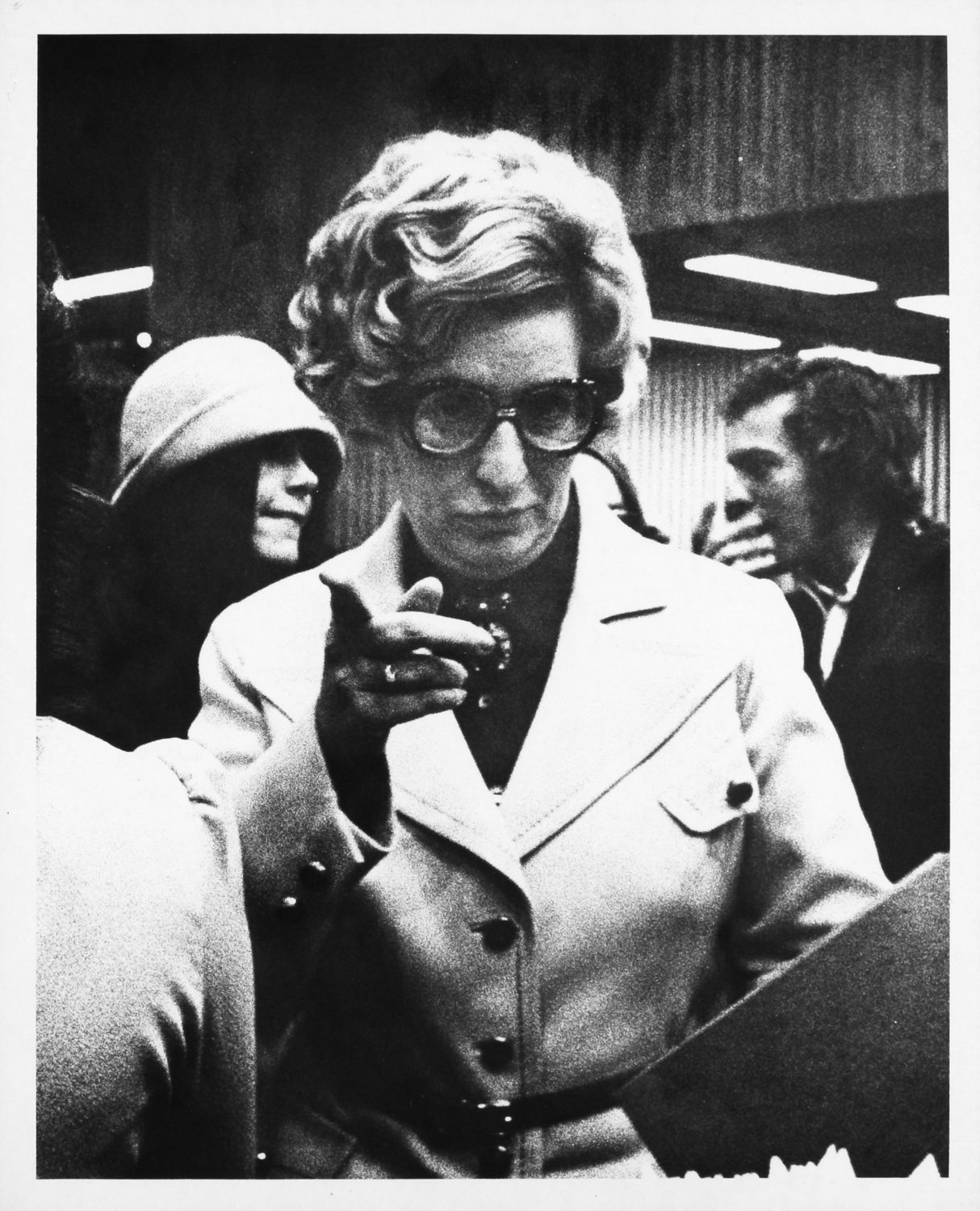 A woman in a coat with glasses pointing at the camera.