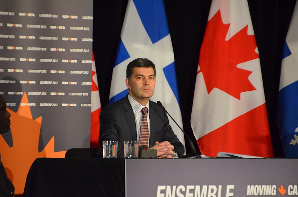 Mario Dumont en 2012 Un homme assis à une table devant un micro. Plusieurs drapeaux canadiens et québécois sont dressés derrière lui.
