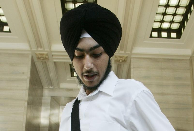 A young man wearing a kirpan, black turban, white shirt, and black pants. He is standing in a tall white room.