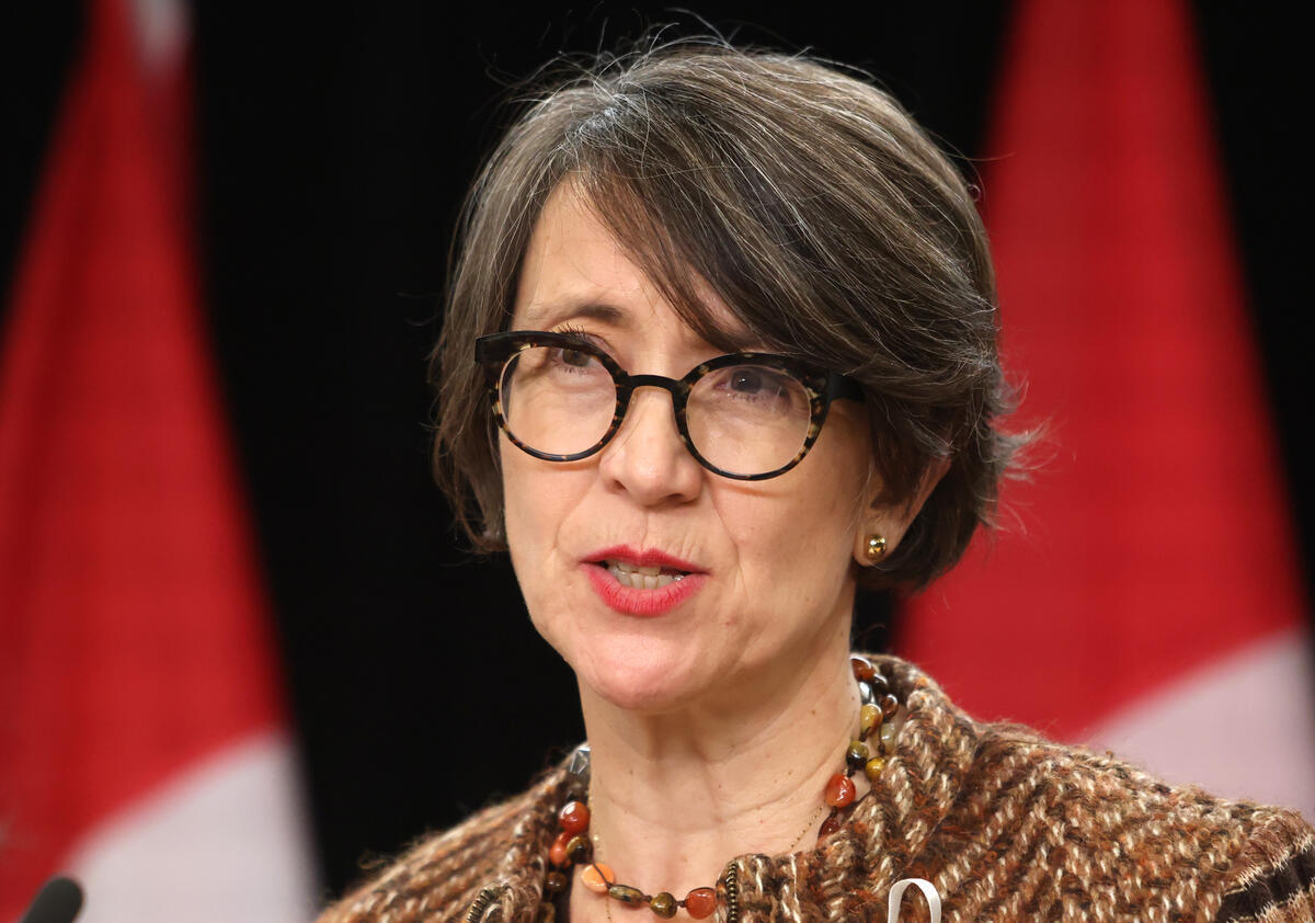 Woman with short hair, glasses, and a coloured necklace. There are two Canadian flags seen behind her.