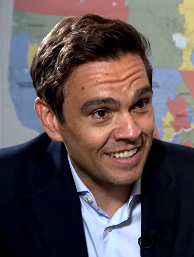 A man in a dark-coloured suit and a light blue shirt. There is a coloured map behind him.
