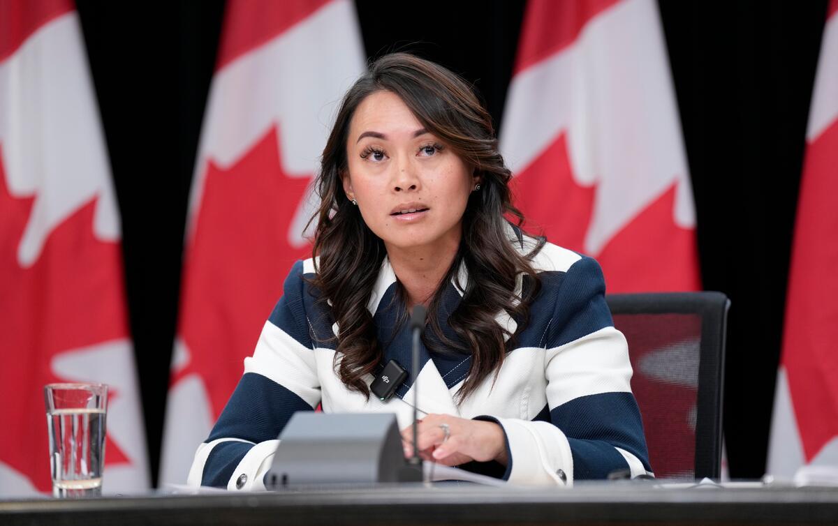 A woman with dark brown hair wearing a striped blue-white outfit is seated at a table with a microphone. Behind her is a row of Canadian flags.