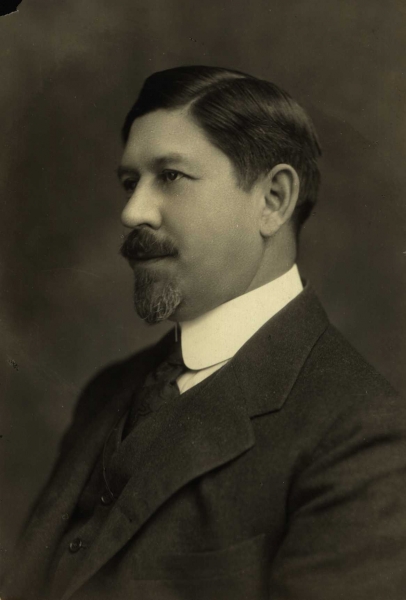 A monochromatic picture of a man in a suit with short hair, a beard, and a mustache.