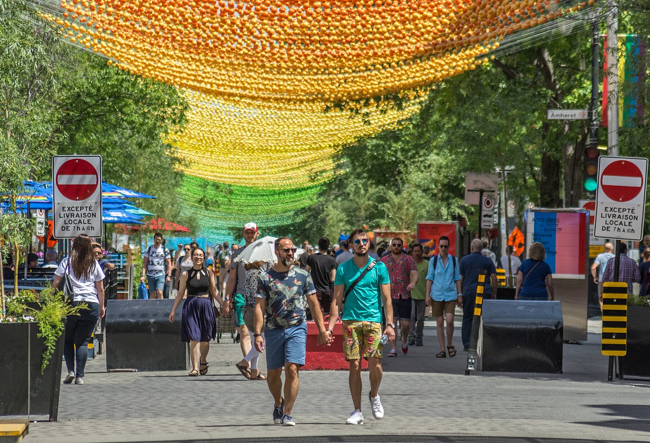 People walking in the street with trees and signs on the side. There are multicoloured balls suspended above.
