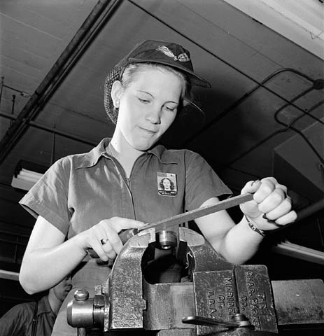 Woman factory worker files a machine part while piped music plays on loudspeakers, November 1943.