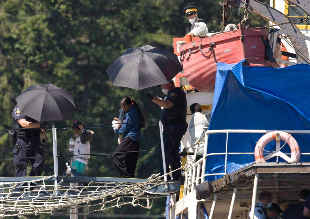 South Asian people escorted off a ship by two CBSA agents holding black umbrellas.