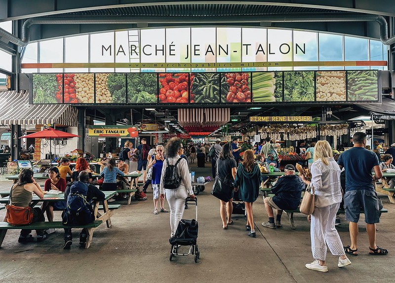 Jean-Talon Market in Montreal A market common area with tables and many people. There's a glass window near the ceiling with the name Marché Jean-Talon written on it.