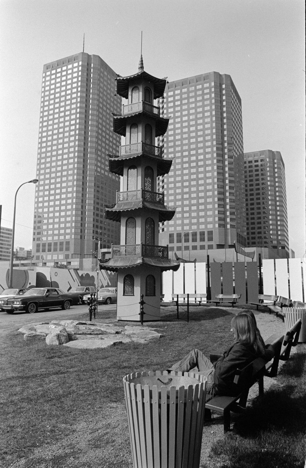 A Chinese pagoda surrounded by benches and a trash bin with tall buildings in the background